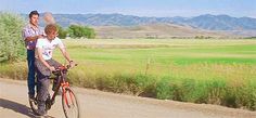 two men riding bikes down a dirt road next to a lush green field with mountains in the background