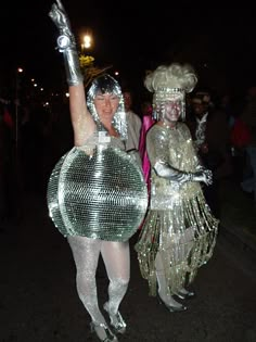 two women dressed in silver and white are walking down the street with their arms up