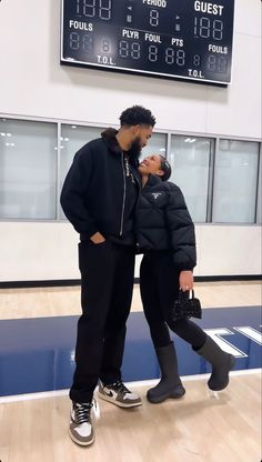 a man and woman standing next to each other in front of a basketball court with the scoreboard behind them