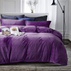 a bed with purple comforter and pillows in a white room next to a window