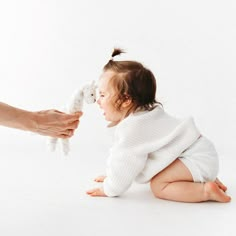 a baby is sitting on the floor and being held by someone's hand with a stuffed animal