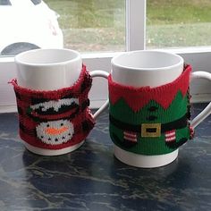 two coffee mugs with knitted christmas sweaters on them sitting next to a window