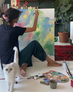 a woman sitting on the floor next to a white cat in front of a painting