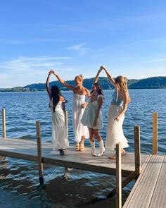 four women in white dresses standing on a dock with their arms up and hands together