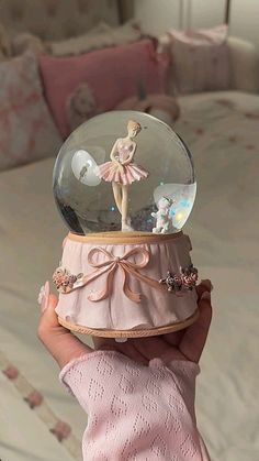 a hand holding a snow globe with a ballerina figure in it, on top of a bed