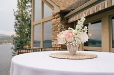 a vase with flowers sitting on top of a table near a building by the water