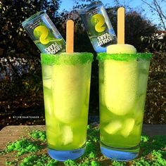 two glasses filled with green liquid and popsicle sticks on top of moss covered ground