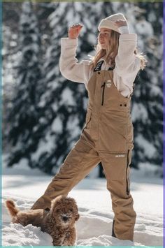 a woman standing in the snow next to a dog wearing overalls and a hat