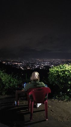 a person sitting on top of a red chair in front of a city at night