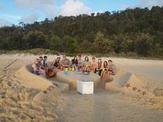 a group of people sitting around a table made out of sand on the beach with trees in the background