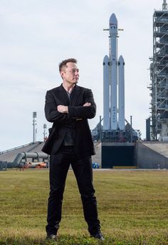 a man standing in the grass with his arms crossed next to a rocket on top of a building