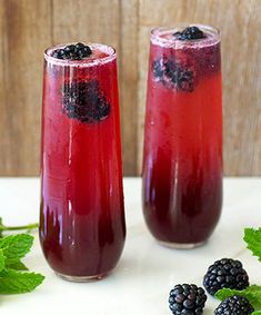 two glasses filled with red liquid and blackberries next to green leaves on a table