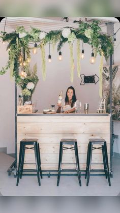 a woman sitting at a bar with three stools in front of her and flowers hanging from the ceiling