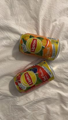 two cans of orange juice sitting on top of a white sheet covered bed next to each other