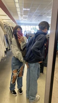 two people standing in front of a mirror looking at their cell phones while wearing ripped jeans