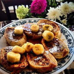 french toast with bananas and powdered sugar on a blue and white plate next to flowers