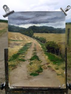 an easel with a painting on it in the middle of a dirt road surrounded by grass and trees