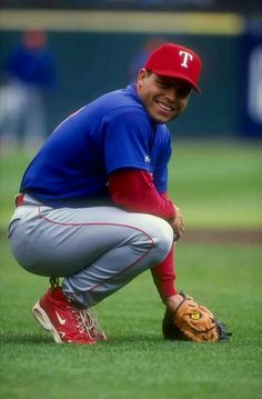 a baseball player squatting on the field with his glove in hand and smiling at the camera