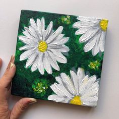 a woman's hand holding up a painted square with white daisies on it