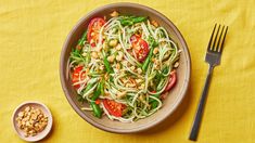 a bowl filled with noodles and vegetables next to a fork on a yellow table cloth