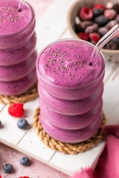 two glasses filled with smoothie on top of a wooden table next to berries and raspberries