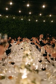 many people are sitting at a long table with wine glasses in front of them and toasting