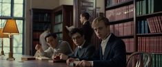 three men sitting at a table with books in front of them and one man holding a book