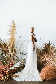 a woman in a white dress standing next to some tall grass and dried plants with her hands on her hips