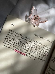 an open book sitting on top of a bed next to a white sheet and a pink flower