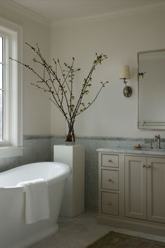 a white bath tub sitting under a window next to a sink and a vase with flowers