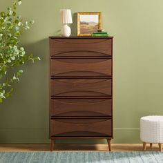 a wooden chest of drawers sitting next to a green wall and a white stool with a plant on top