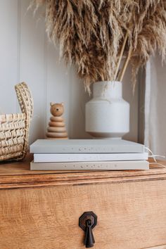 a stack of books sitting on top of a wooden dresser next to a vase with dry grass