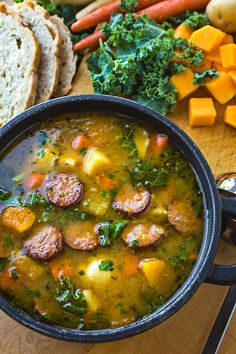 a bowl of soup with sausage, potatoes and carrots on a cutting board next to bread