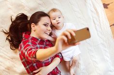 a woman holding a baby and taking a selfie with her cell phone while laying on a bed