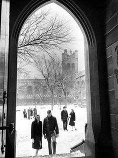 black and white photograph of people walking in the snow through an archway to a building