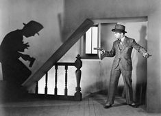 a man in a suit and hat standing next to a stair case with his shadow on the wall