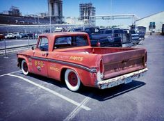 an old red pick up truck parked in a parking lot with other trucks behind it