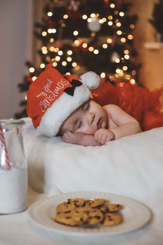 a baby laying on a bed next to a plate of cookies and milk with a christmas tree in the background