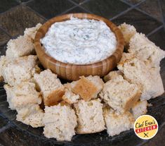 a wooden bowl filled with white dip surrounded by bread cubes on a glass plate