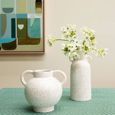 two white vases sitting on top of a table with flowers in them and a painting behind them