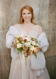 a woman in a wedding dress holding a bouquet and fur stole around her waist, smiling at the camera