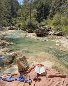 an open book sitting on top of a blanket next to a river in the woods