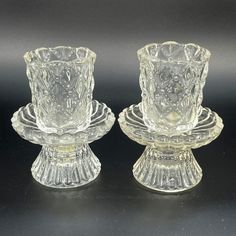 two clear glass candlesticks sitting side by side on a black surface with one candle holder in the middle