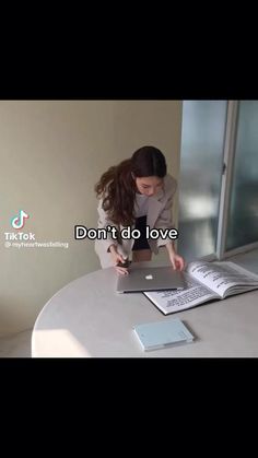 a woman leaning over a book on top of a table next to an open laptop computer