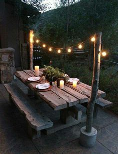 a wooden table with candles and plates on it in front of some lights hanging from the trees