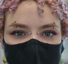 a woman with pink hair wearing a black face mask and gold studs on her eyes