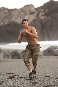 a shirtless man holding a football on the beach