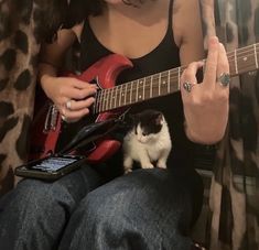 a woman playing guitar with a cat sitting on her lap next to her and looking at the camera