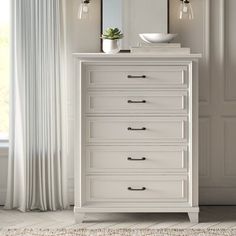 a white chest of drawers with a mirror and bowl on top, in front of a window