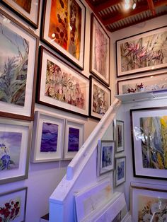 many framed pictures hang on the wall next to a stair case in a room filled with paintings